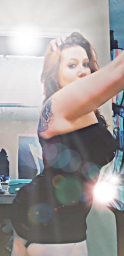 Curvy woman with dark hair and a visible tattoo on her arm, wearing a black top and white bottoms, posing indoors with