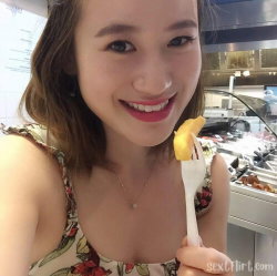 A smiling woman with shoulder-length brown hair wears a floral top and holds a fork with food, taking a selfie in a