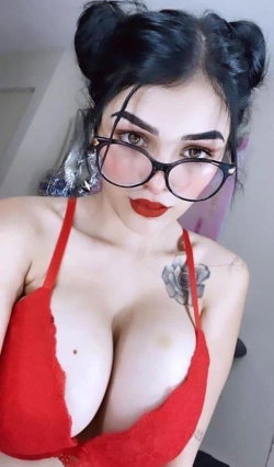 Woman with dark hair in twin buns, wearing black glasses and a red lace bra, posing indoors with visible shoulder