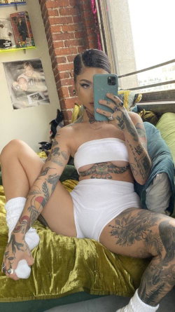 A tattooed woman with dark hair takes a mirror selfie in white lingerie, sitting on a green couch near a brick wall.