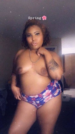Curvy woman with curly hair poses topless in floral bikini bottoms, hand on chin, in a dimly lit room.