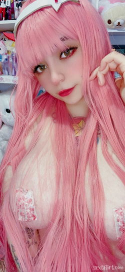 Person with long pink wig and white headband, wearing floral pasties, posing in a room with plush toys and shelves.