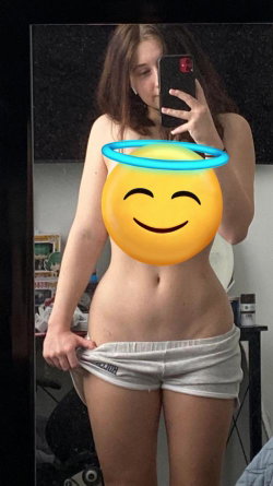 A young woman with shoulder-length brown hair takes a mirror selfie, wearing low-rise white shorts and holding her