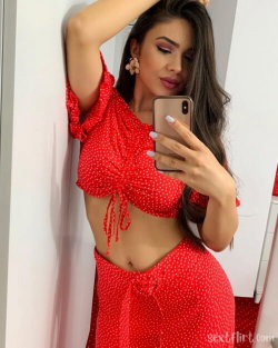 Brunette woman in red polka-dot crop top and matching skirt takes a mirror selfie, hand in hair, posing against a white