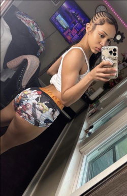 A woman with braided hair takes a mirror selfie in a white tank top and colorful patterned shorts, showing a toned