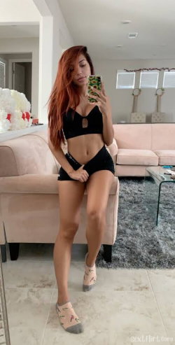 A woman with long red hair in a black crop top and shorts takes a mirror selfie in a living room, wearing patterned