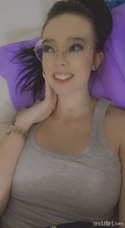 A woman with dark hair and round glasses lies on a purple pillow, wearing a gray tank top and smiling at the camera.