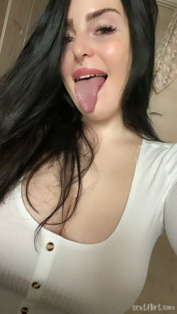 A woman with long dark hair and a nose ring sticks her tongue out playfully in a selfie, wearing a white button-up top