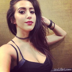 Brunette woman with long dark hair wearing a black lace top and choker, posing indoors with hand in hair.