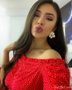 Brunette woman in a red polka-dot off-shoulder top making a playful kiss face, indoors with soft lighting.