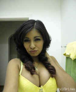 Brunette woman in a yellow lace top poses in a bathroom, hand on neck, looking at the camera.