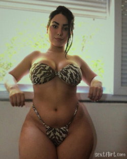 Curvy woman in a patterned bikini poses indoors near a window, looking at the camera with hands on the sill.