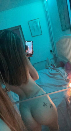 A woman with shoulder-length hair takes a mirror selfie in a dimly lit bedroom, showing her bare back and buttocks with