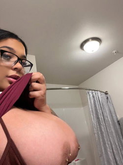 A woman with dark hair and glasses in a bathroom, pulling down a maroon top to reveal her bare breast with nipple