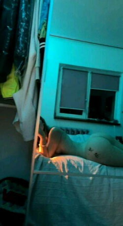 A person with a curvy build lies on a bed in a dimly lit room, partially covered by sheets, with visible tattoos on the