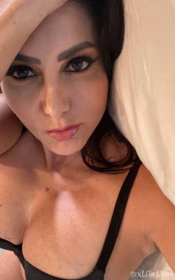Brunette woman with defined features in black lingerie, partially covered by a sheet, taking a close-up selfie indoors.