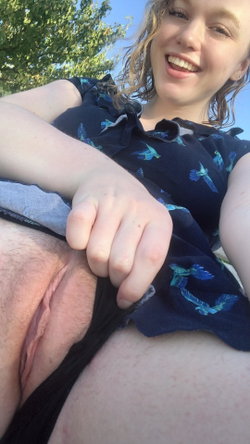 A smiling woman with light brown hair in a blue patterned top, outdoors, holding her lap with one hand.