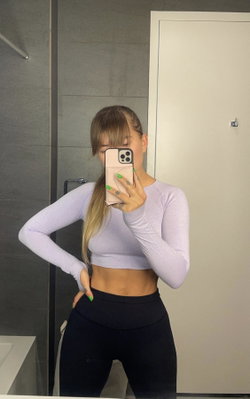 A slim woman with long blonde hair takes a mirror selfie in a lavender crop top and black leggings, standing in a
