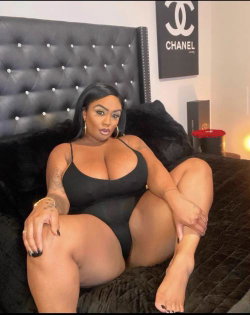 Curvy woman with long dark hair sits on a black bed in a black bodysuit, posing with one leg bent and hand on thigh.