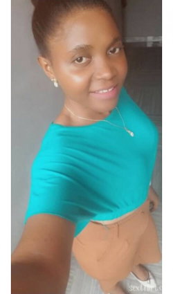 A woman with dark skin and short hair wears a teal top and tan skirt, smiling in a selfie pose indoors.