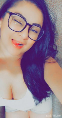 A woman with long dark hair and glasses smiles playfully in a close-up selfie, wearing a white top while lying down.