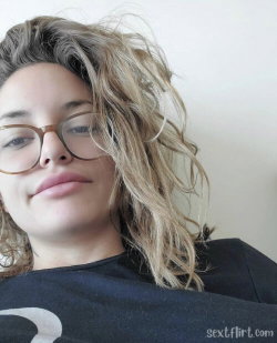 A woman with wavy blonde hair and glasses, wearing a black top, resting against a plain background in a close-up selfie.
