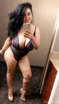 Curvy woman with long dark hair takes mirror selfie in black lace bodysuit and high heels in a hotel room.