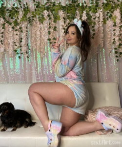 Curvy woman in tie-dye dress and pastel Crocs poses on a white couch with a small dog, surrounded by floral decor.