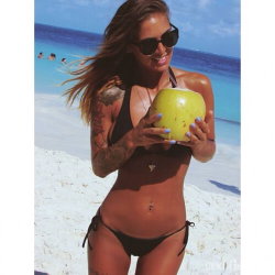Brunette woman in black bikini holding a coconut on a sunny beach, with visible tattoos on her arms and legs.