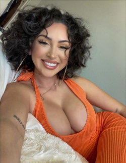 A smiling woman with curly dark hair and a nose ring wears an orange ribbed halter top, lying on a white fluffy surface