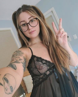 A woman with long blonde hair and glasses takes a selfie in a black lace top, showing arm tattoos and making a peace