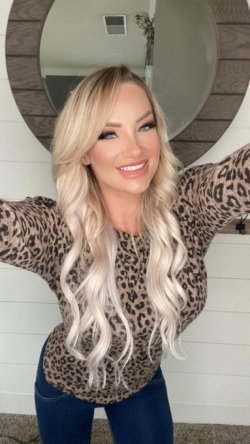 Blonde woman with wavy hair in a leopard print top and jeans, posing in front of a round mirror with arms outstretched.