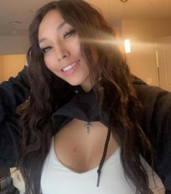 A woman with long dark wavy hair smiles in a selfie, wearing a white top under a black hoodie in a warmly lit indoor