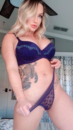 Blonde woman in purple lace lingerie poses in a bedroom, showcasing a curvy figure with visible tattoos on her side and