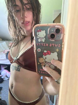 A woman with wet hair takes a mirror selfie in a brown lace-trim bra and matching underwear, showing a tattoo on her