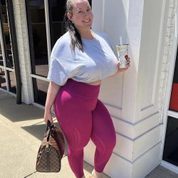 A curvy woman in a loose white top and tight magenta leggings stands outdoors, holding a drink and a patterned bag,