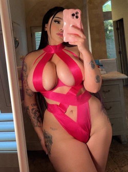 A woman with long dark hair and multiple tattoos takes a mirror selfie in a bright pink cutout bodysuit, standing in a