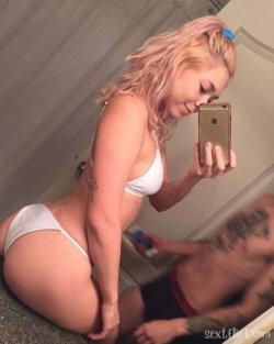 A woman with pink hair in a white bikini takes a mirror selfie while kneeling, with a tattooed man partially visible