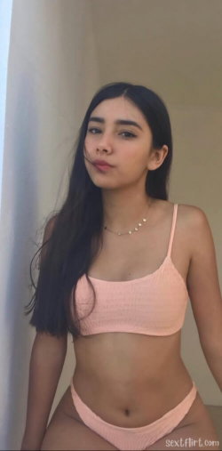 A young woman with long dark hair wears a peach-colored bikini, posing indoors near a wall.