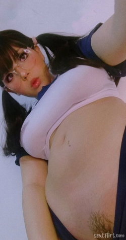 A woman with dark hair in pigtails and glasses wears a cropped pink top and blue shorts, posing playfully with her arm