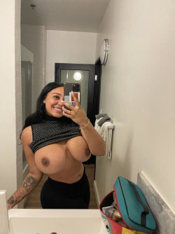 A woman with long dark hair and a tattooed arm takes a mirror selfie in a bathroom, wearing a black top and bottoms,