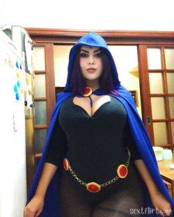 A woman in a black bodysuit and blue hooded cape poses indoors, wearing a gold chain belt with red accents.