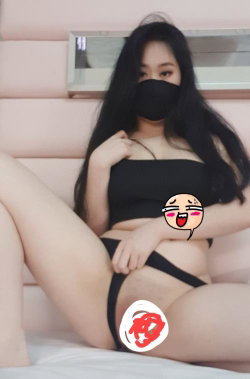 A woman with long dark hair sits on a bed wearing a black strapless top and black thong underwear, covering her face