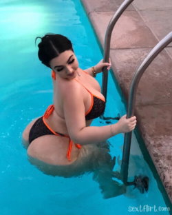 Curvy woman in black and orange bikini posing in a swimming pool, holding ladder with one hand, looking over shoulder.