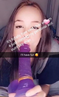 Brunette woman with long hair in a close-up selfie, holding a purple toy to her mouth while lying down indoors.