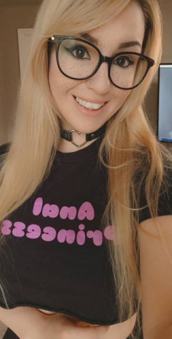 Blonde woman with glasses and a black crop top, smiling in a selfie-style photo indoors.