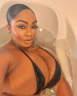 A curvy Black woman with dark hair and bold makeup poses in a bubble bath wearing a black string bikini top.