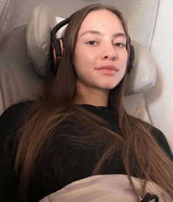 A young woman with long brown hair wearing black headphones and a dark top, reclining on a light-colored seat.