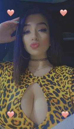 Woman with dark hair and makeup wearing a yellow leopard-print top with a deep neckline, posing for a selfie with heart