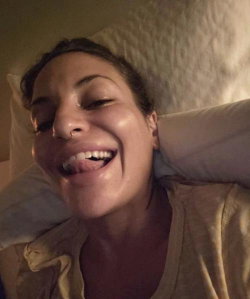 A woman with dark hair and a nose ring smiles broadly while lying in bed, wearing a light-colored top.